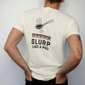 Man wearing a Ivory tee, seen from the back: a cupping spoon at a cartoon mouth mid-slurp over a row of cupping bowls, with Slurp in caps and Like A Pro below, drawn in a single fine crema-cream line with one burnt copper accent.