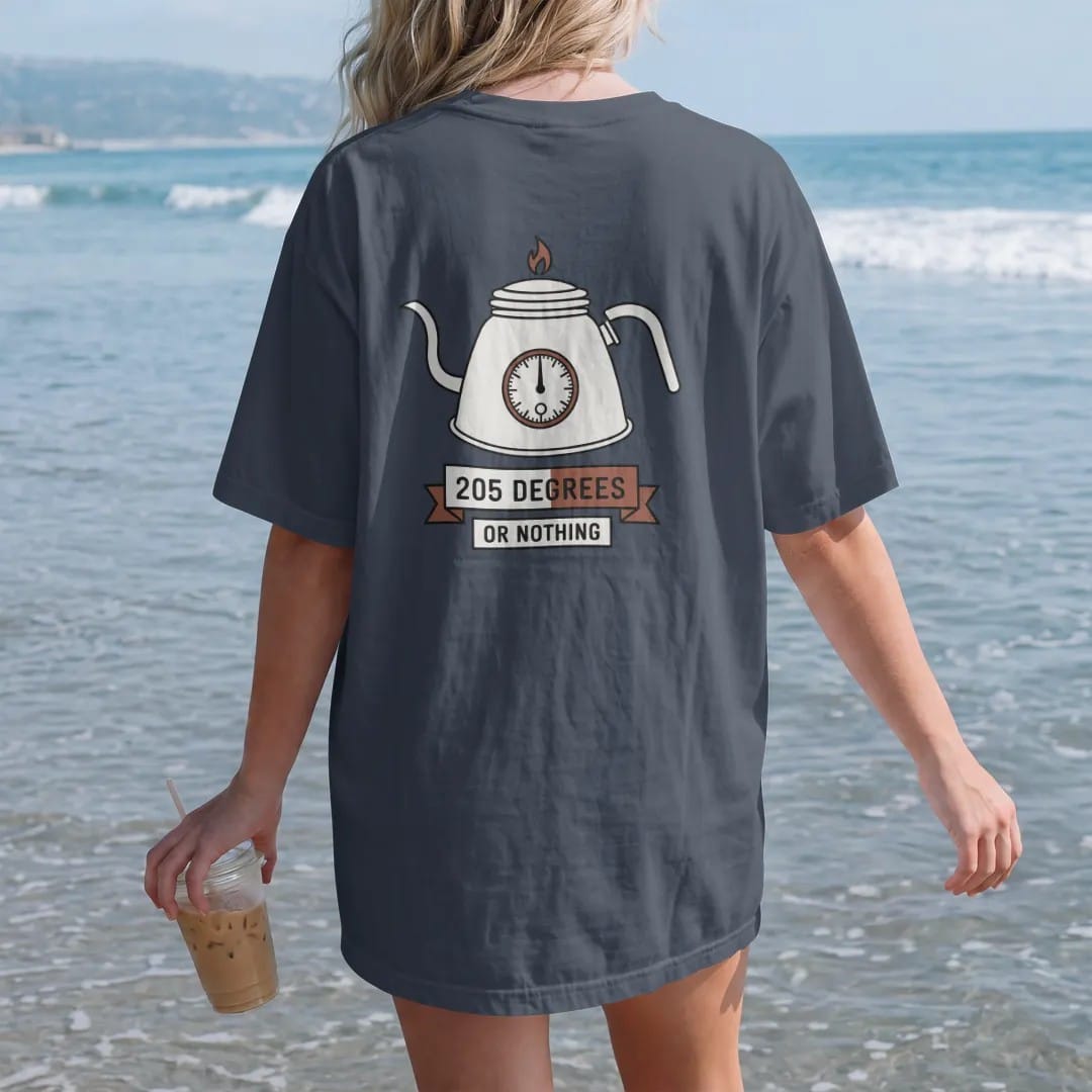 Woman on the beach in a Midnight tee, back view: a gooseneck kettle with a dial thermometer on its lid, 205 Degrees in clean numerals and Or Nothing on a banner, drawn in a single fine crema-cream line with one burnt copper accent.