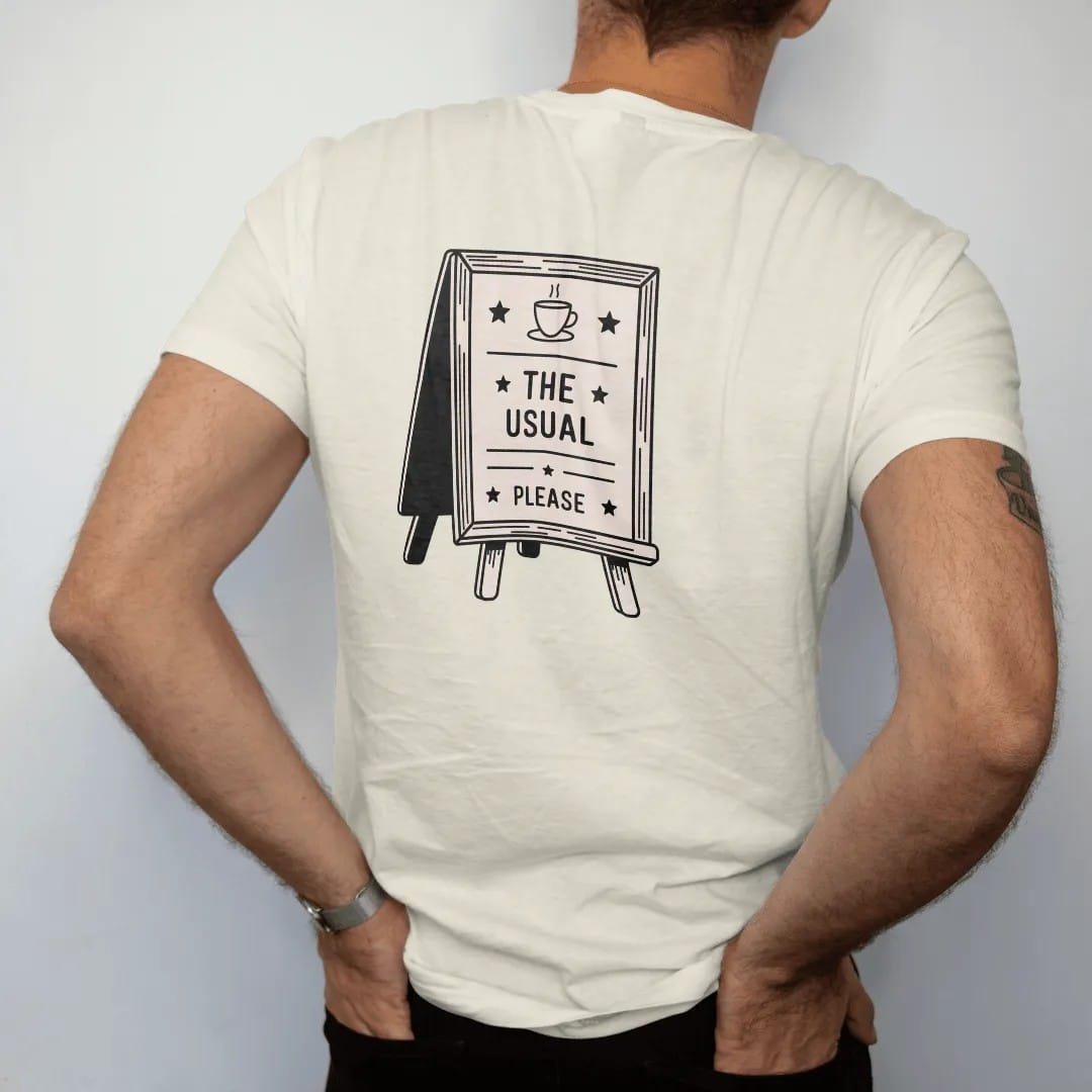 Man wearing a Ivory tee, seen from the back: a café letter board on an easel topped with a coffee cup icon, reading The Usual with Please underneath, drawn in a single fine roast-brown line with one burnt copper accent.
