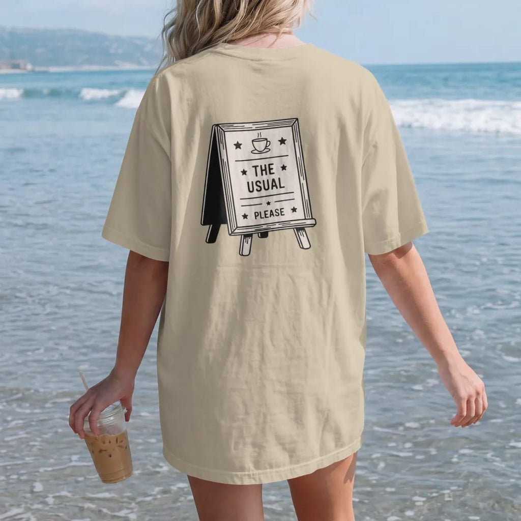 Woman on the beach in a Sandstone tee, back view: a café letter board on an easel topped with a coffee cup icon, reading The Usual with Please underneath, drawn in a single fine roast-brown line with one burnt copper accent.