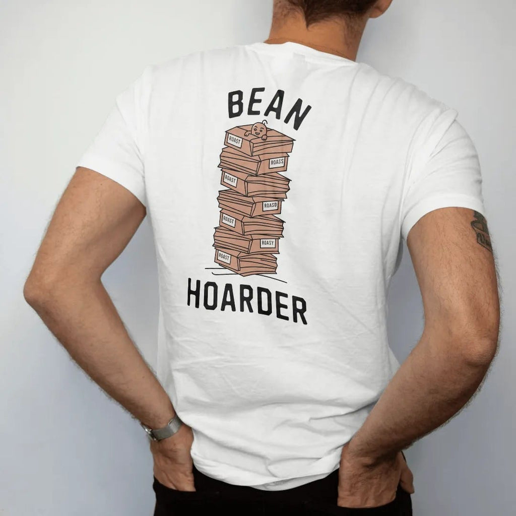Man wearing a White tee, seen from the back: a wobbly stack of labeled coffee bags with the bean mascot peeking out, Bean above and Hoarder below, drawn in a single fine crema-cream line with one burnt copper accent.