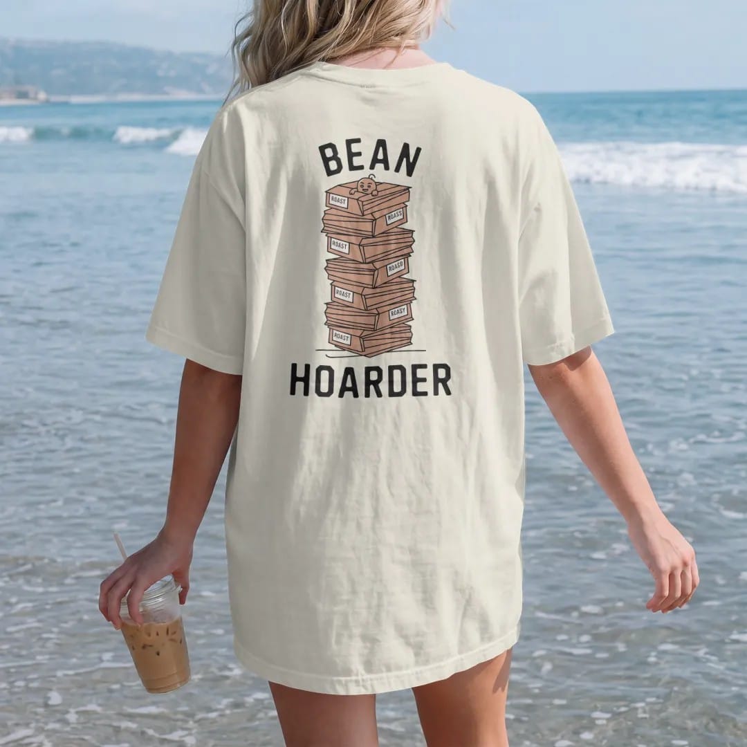 Woman on the beach in a Ivory tee, back view: a wobbly stack of labeled coffee bags with the bean mascot peeking out, Bean above and Hoarder below, drawn in a single fine crema-cream line with one burnt copper accent.