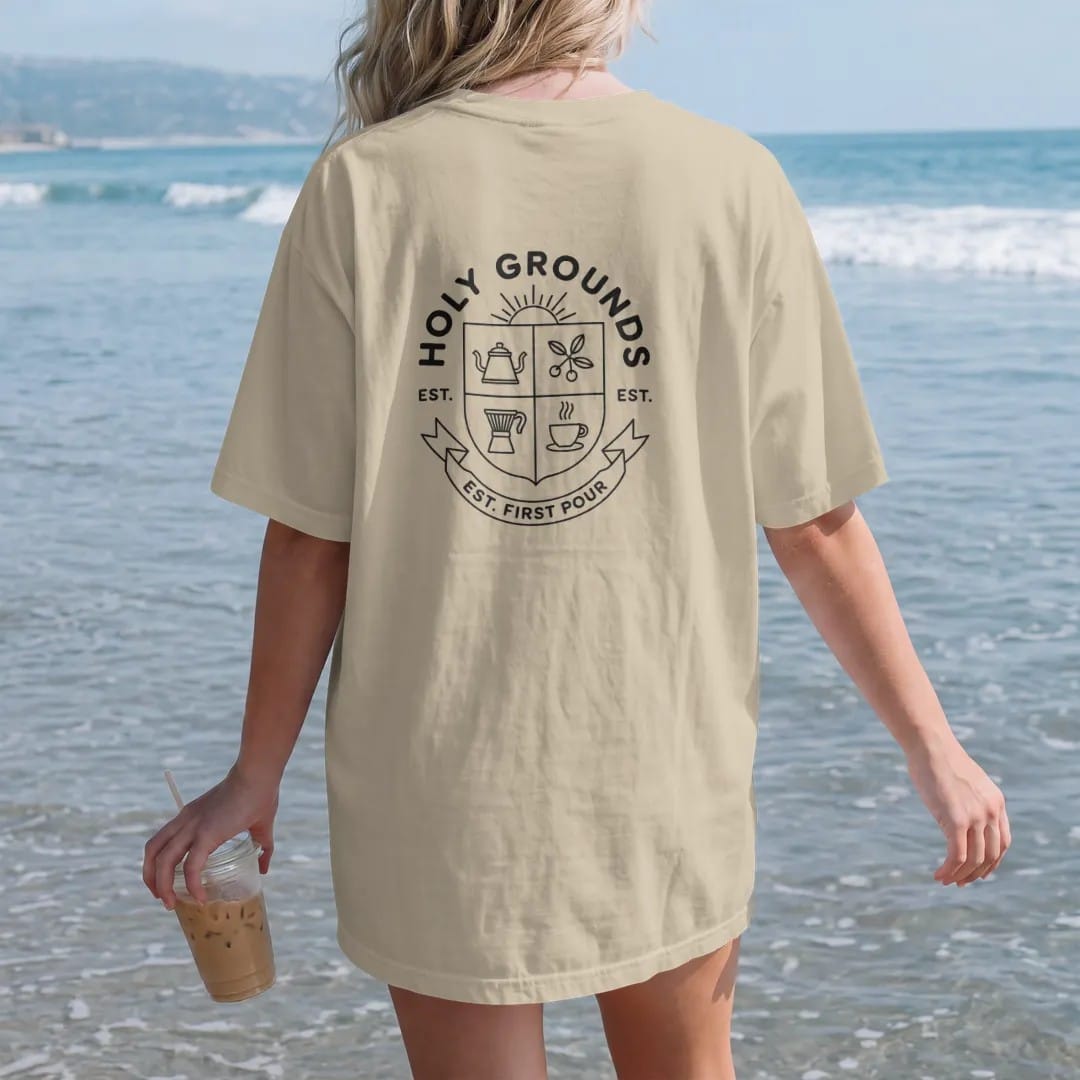 Woman on the beach in a Sandstone tee, back view: a quartered crest with a kettle, portafilter, coffee branch and cup under a steam halo, Holy Grounds arched above and Est. First Pour on a ribbon, drawn in a single fine crema-cream line with one…