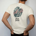 Man wearing a Ivory tee, seen from the back: a single large monstera leaf with split fenestrations, small brush-written caps "PLANT PARENT" beneath, painted in loose watercolor and ink with soft deep green, terracotta, soil brown and moss washes.