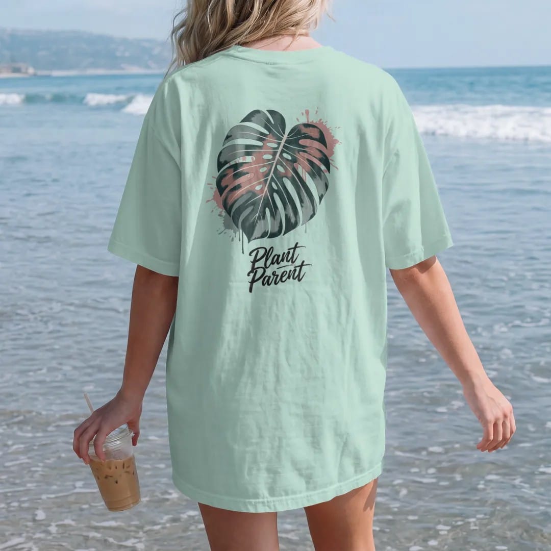 Woman on the beach in a Chalky Mint tee, back view: a single large monstera leaf with split fenestrations, small brush-written caps "PLANT PARENT" beneath, painted in loose watercolor and ink with soft deep green, terracotta, soil brown and moss…