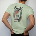 Man wearing a Light Green tee, seen from the back: a simple window with soft angled light rays falling on a small potted plant, "BRIGHT" in loose brush script above and "INDIRECT LIGHT" small below, painted in loose watercolor and ink with soft…