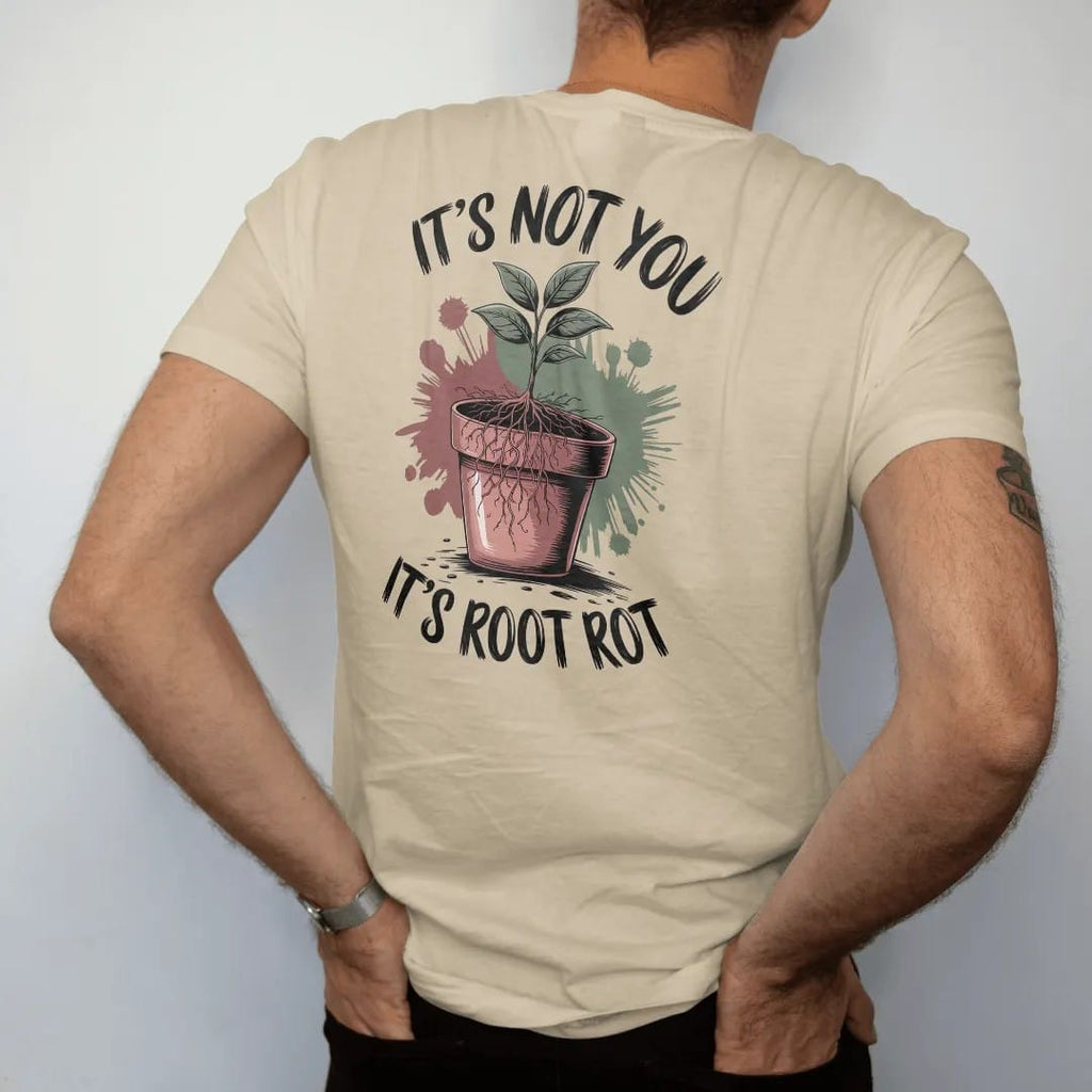 Man wearing a Sandstone tee, seen from the back: a small plant lifted out of its pot with tangled brown roots showing, "IT'S NOT YOU" in loose brush script above and "IT'S ROOT ROT" below, painted in loose watercolor and ink with soft deep green,…