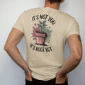Man wearing a Sandstone tee, seen from the back: a small plant lifted out of its pot with tangled brown roots showing, "IT'S NOT YOU" in loose brush script above and "IT'S ROOT ROT" below, painted in loose watercolor and ink with soft deep green,…