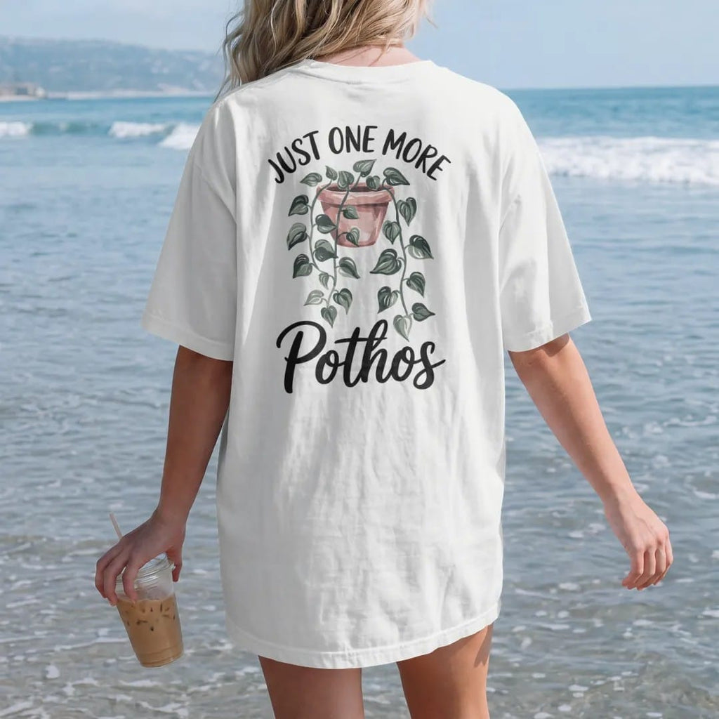 Woman on the beach in a White tee, back view: a trailing pothos vine draping from a small hanging pot with heart-shaped leaves, "JUST ONE MORE" in small brush-written caps above and "POTHOS" in loose brush script below, painted in loose watercolor…