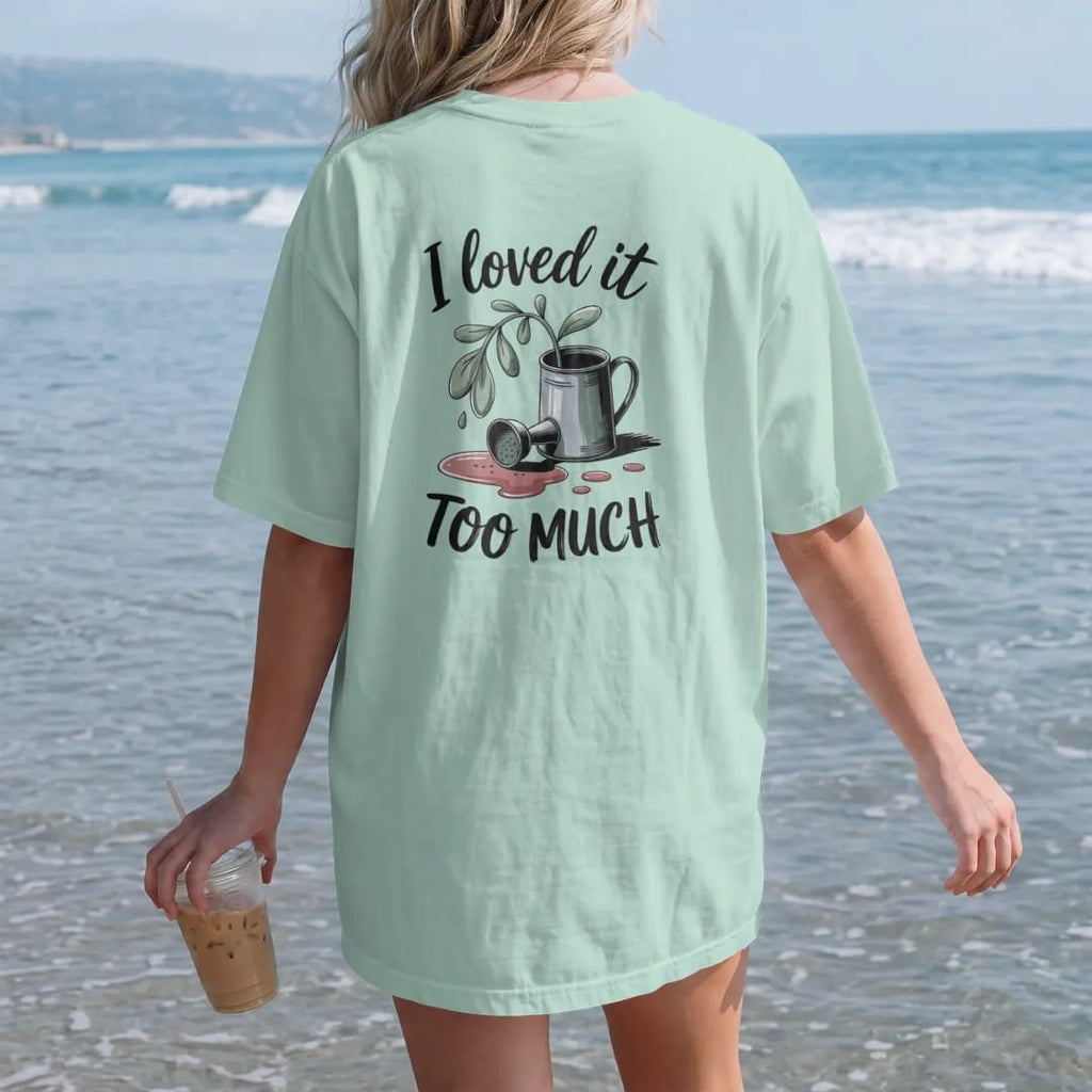 Woman on the beach in a Chalky Mint tee, back view: a small droopy plant beside a tipped-over watering can with a puddle, "I LOVED IT" in loose brush script above and "TOO MUCH" in small brush-written caps below, painted in loose watercolor and ink…