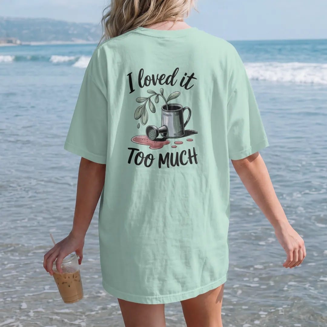 Woman on the beach in a Chalky Mint tee, back view: a small droopy plant beside a tipped-over watering can with a puddle, "I LOVED IT" in loose brush script above and "TOO MUCH" in small brush-written caps below, painted in loose watercolor and ink…