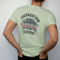 Man wearing a Light Green tee, seen from the back: a row of three small glass jars with rooting cuttings on a thin shelf, "PROPAGATION" arched above and "STATION" small below in small brush-written caps, painted in loose watercolor and ink with…