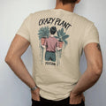 Man wearing a Sandstone tee, seen from the back: a figure holding an armful of potted plants, "CRAZY PLANT" in loose brush script above and "PERSON" small below, painted in loose watercolor and ink with soft deep green, terracotta, soil brown and…