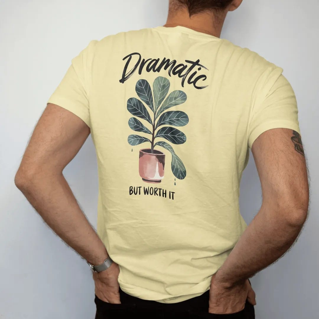 Man wearing a Butter tee, seen from the back: a fiddle leaf fig in a tall pot with one leaf dropping dramatically, "DRAMATIC" in loose brush script above and "BUT WORTH IT" small below, painted in loose watercolor and ink with soft deep green,…