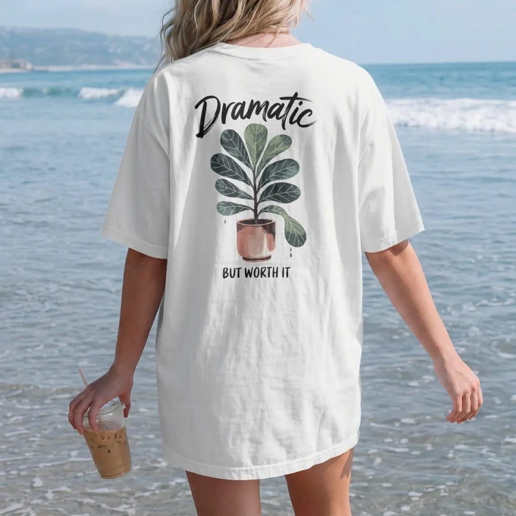 Woman on the beach in a White tee, back view: a fiddle leaf fig in a tall pot with one leaf dropping dramatically, "DRAMATIC" in loose brush script above and "BUT WORTH IT" small below, painted in loose watercolor and ink with soft deep green,…