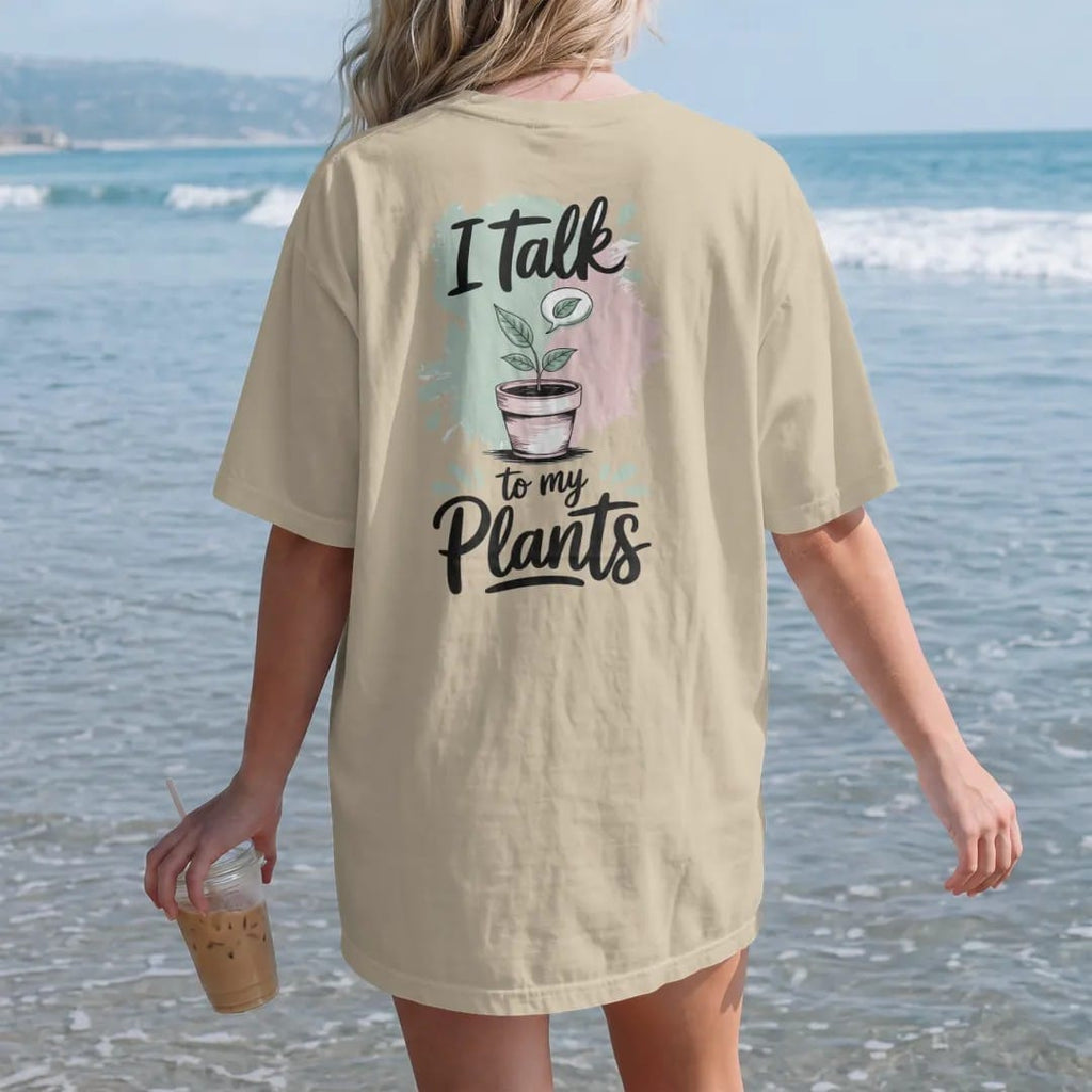 Woman on the beach in a Sandstone tee, back view: a small potted plant with a tiny speech bubble holding a single leaf, "I TALK" in loose brush script above and "TO MY PLANTS" below, painted in loose watercolor and ink with soft mint, clay pink,…