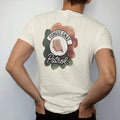 Man wearing a Ivory tee, seen from the back: a small round badge with a sticky trap card and a tiny cartoon gnat, "FUNGUS GNAT" arched above and "PATROL" below in loose brush script, painted in loose watercolor and ink with soft deep green,…