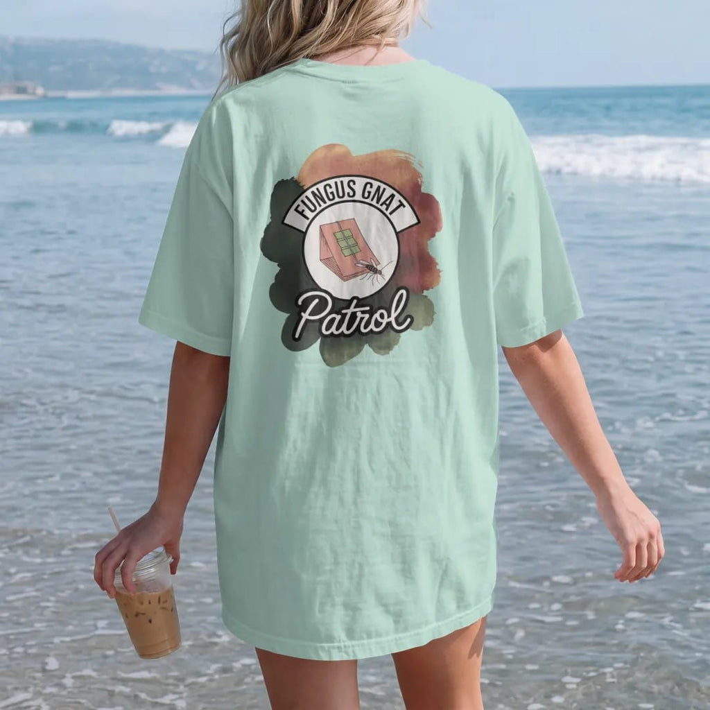 Woman on the beach in a Chalky Mint tee, back view: a small round badge with a sticky trap card and a tiny cartoon gnat, "FUNGUS GNAT" arched above and "PATROL" below in loose brush script, painted in loose watercolor and ink with soft deep green,…