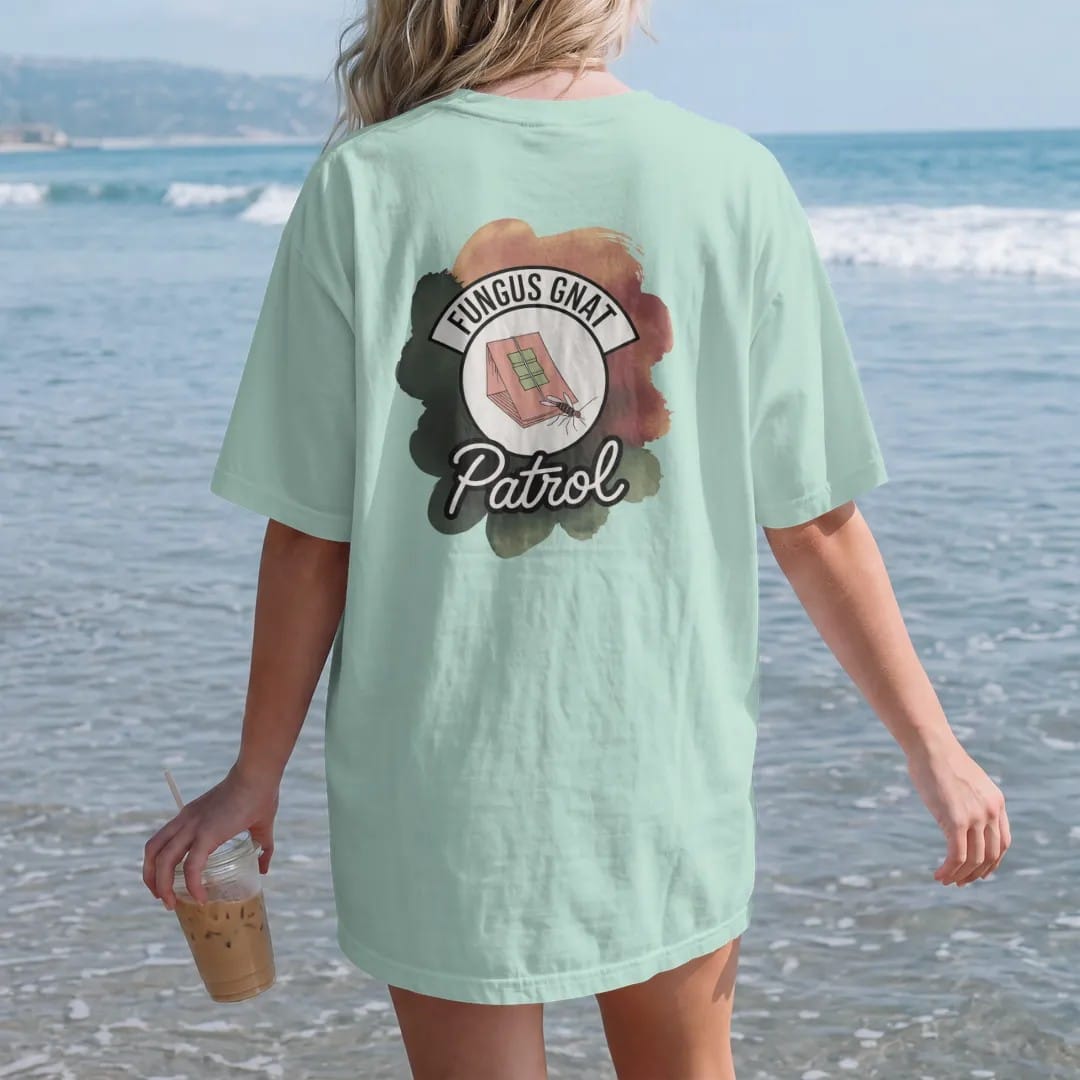Woman on the beach in a Chalky Mint tee, back view: a small round badge with a sticky trap card and a tiny cartoon gnat, "FUNGUS GNAT" arched above and "PATROL" below in loose brush script, painted in loose watercolor and ink with soft deep green,…