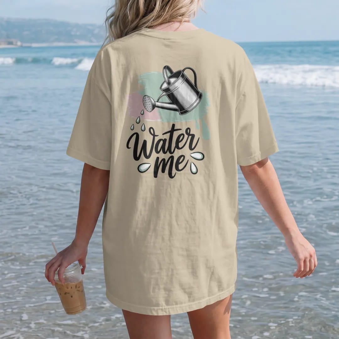 Woman on the beach in a Sandstone tee, back view: a vintage watering can tilted mid-pour with three clean drops, "WATER ME" in hand-lettered brush script beneath, painted in loose watercolor and ink with soft mint, clay pink, cream and lime washes.