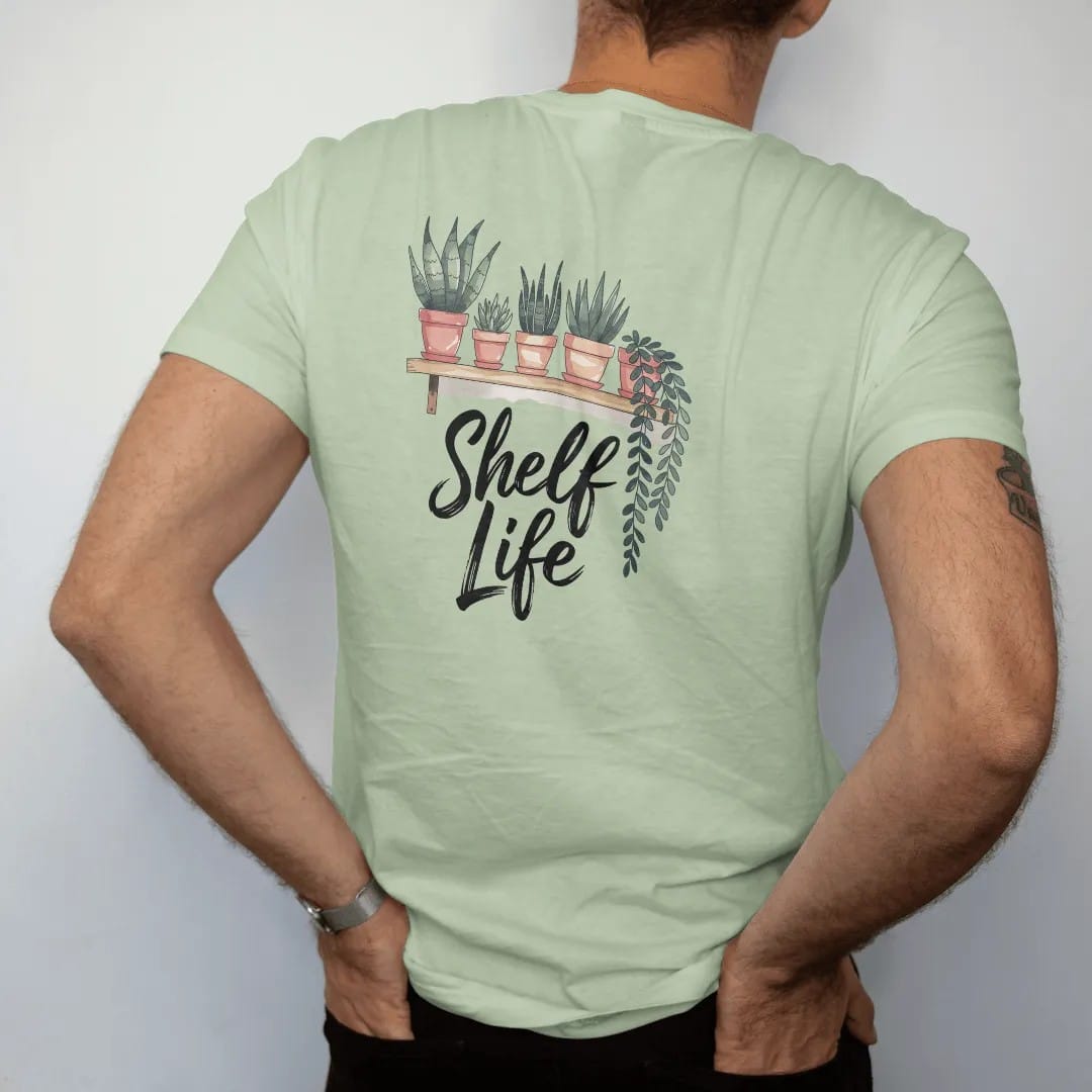 Man wearing a Light Green tee, seen from the back: a minimal floating shelf holding four small pots with different plants, trailing leaves hanging over the edge, "SHELF LIFE" in loose brush script beneath, painted in loose watercolor and ink with…
