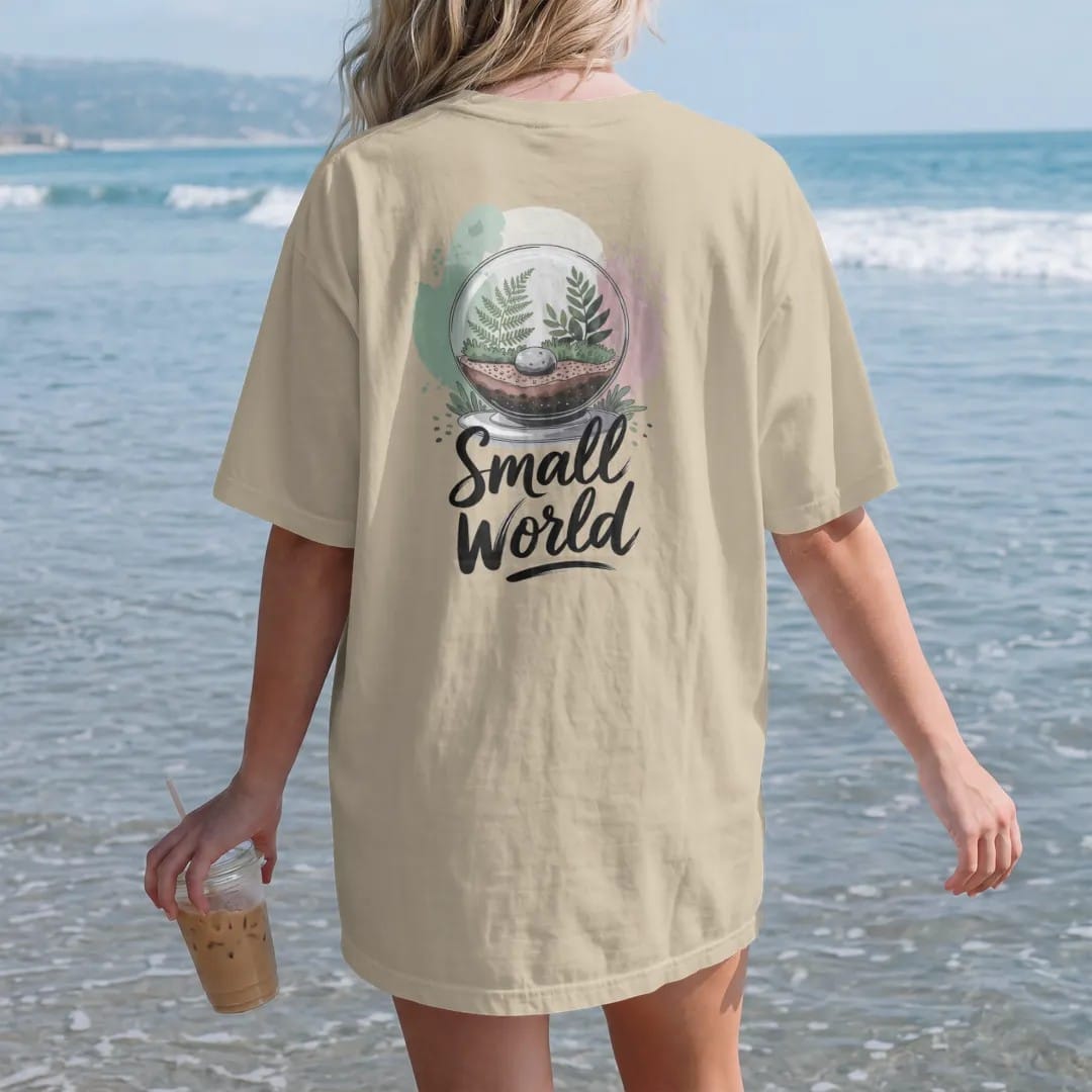 Woman on the beach in a Sandstone tee, back view: a glass globe terrarium with tiny ferns, moss and a pebble inside, "SMALL WORLD" in loose brush script beneath, painted in loose watercolor and ink with soft mint, clay pink, cream and lime washes.