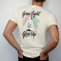 Man wearing a Ivory tee, seen from the back: a small plant under a hanging grow lamp with soft light rays, "GROW LIGHT" in loose brush script above and "GLOW UP" in hand-lettered brush script below, painted in loose watercolor and ink with soft…