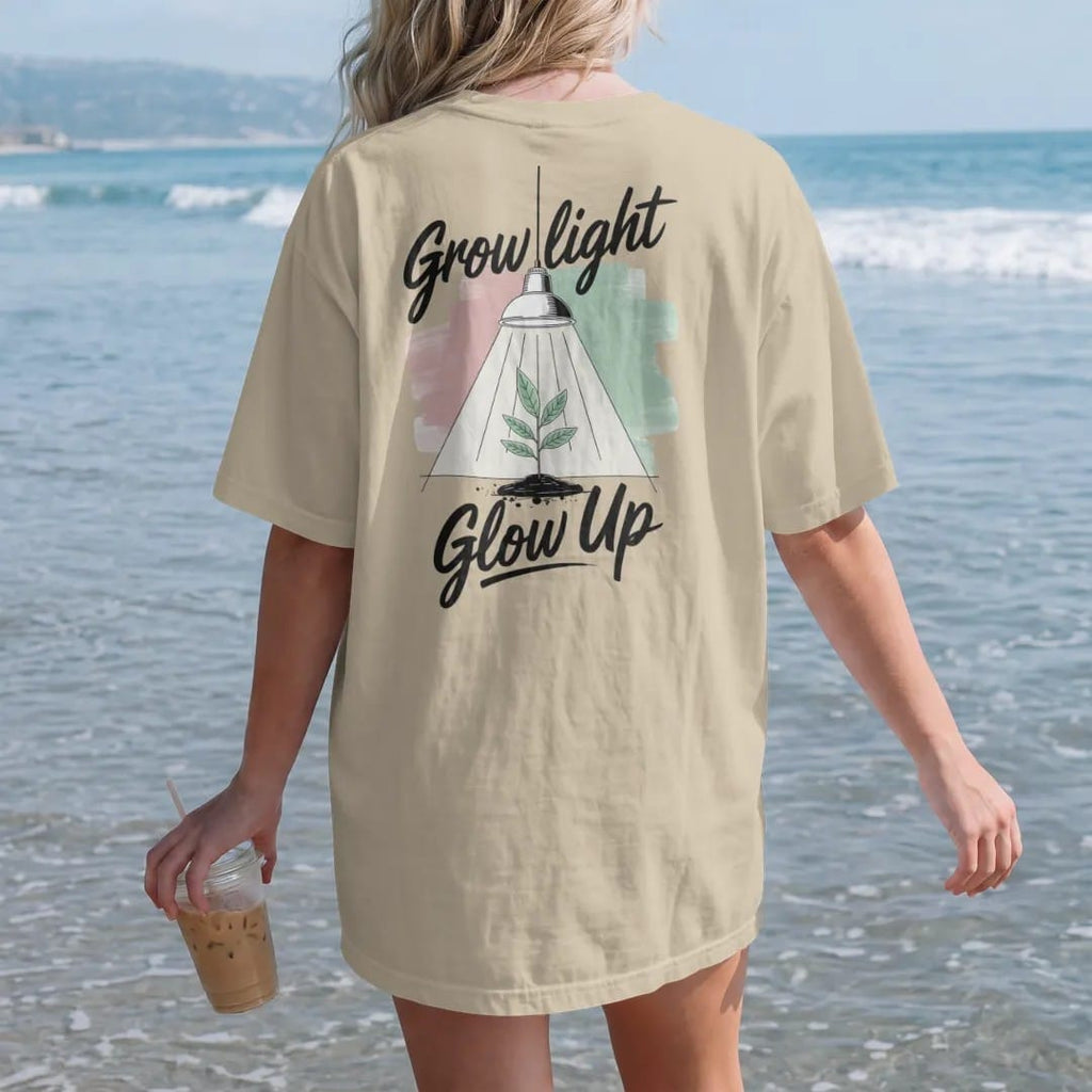 Woman on the beach in a Sandstone tee, back view: a small plant under a hanging grow lamp with soft light rays, "GROW LIGHT" in loose brush script above and "GLOW UP" in hand-lettered brush script below, painted in loose watercolor and ink with…