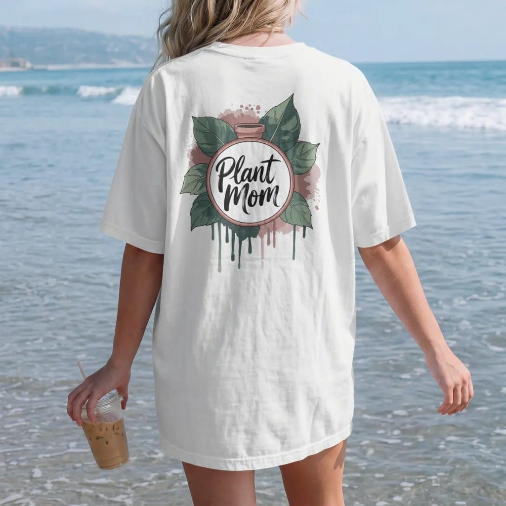Woman on the beach in a White tee, back view: a neat round badge framed by two pothos leaves with a small pot in the center, "PLANT MOM" in loose brush script across the bottom, painted in loose watercolor and ink with soft deep green, terracotta,…