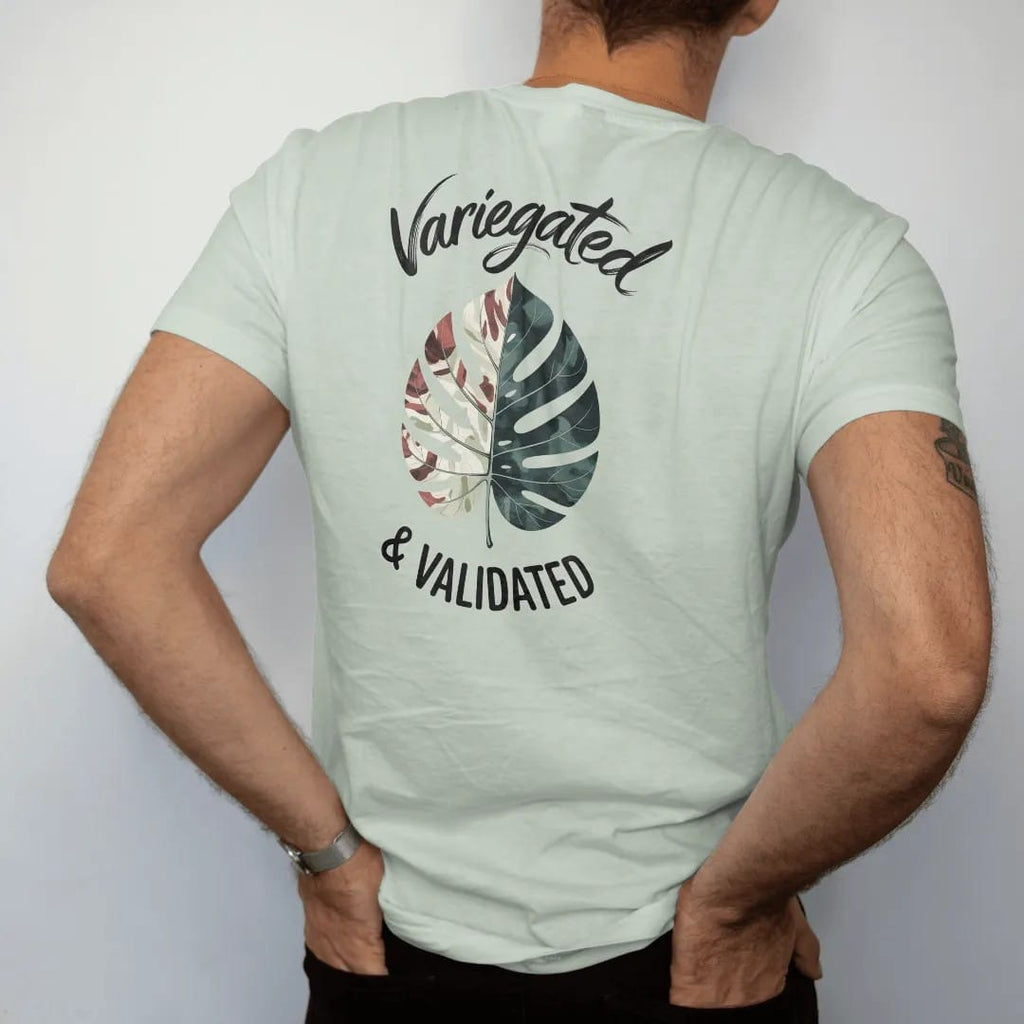 Man wearing a Bay tee, seen from the back: a single variegated leaf with marbled cream and green patches, "VARIEGATED" in loose brush script above and "& VALIDATED" small below, painted in loose watercolor and ink with soft deep green, terracotta,…