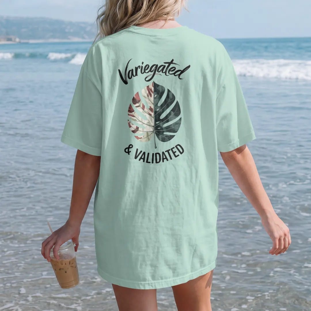 Woman on the beach in a Chalky Mint tee, back view: a single variegated leaf with marbled cream and green patches, "VARIEGATED" in loose brush script above and "& VALIDATED" small below, painted in loose watercolor and ink with soft deep green,…