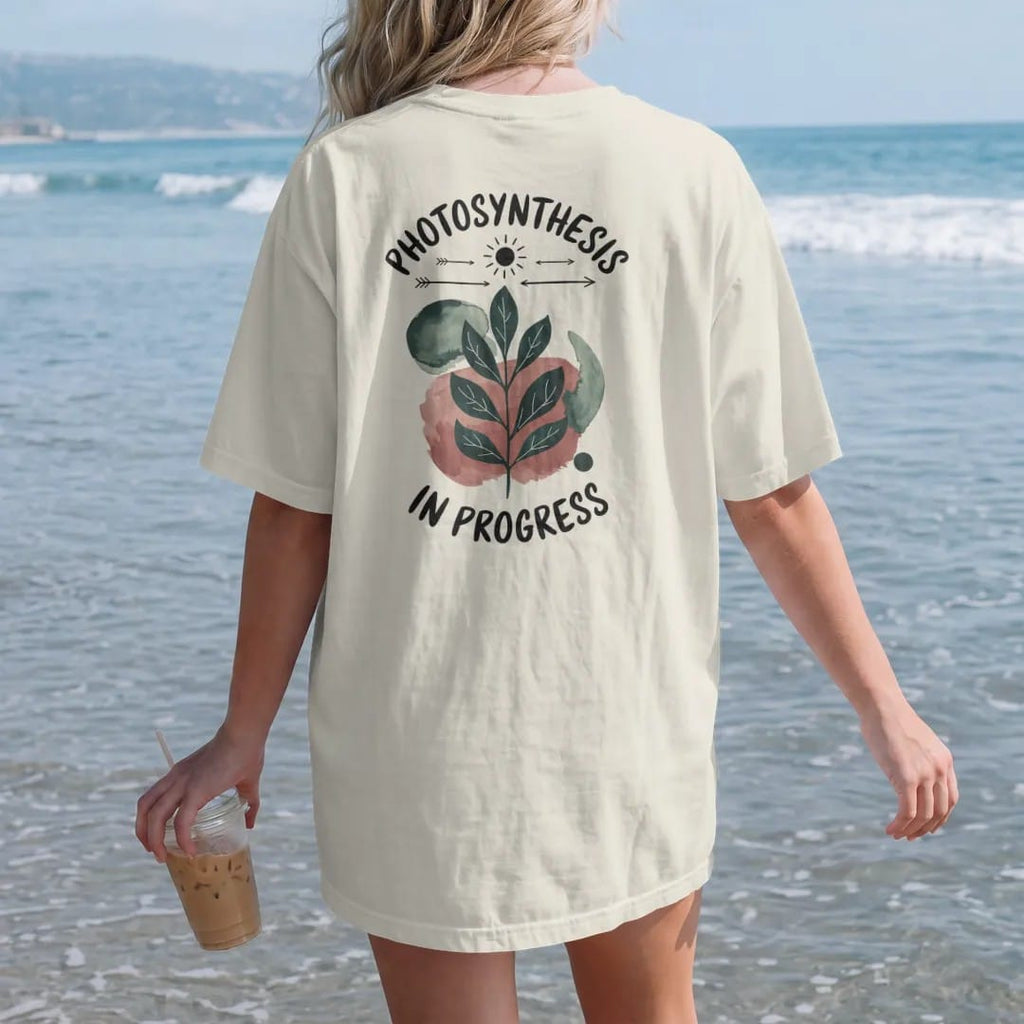 Woman on the beach in a Ivory tee, back view: a single leaf with simple veins and a small sun above it, thin arrows showing light in, "PHOTOSYNTHESIS" in small brush-written caps above and "IN PROGRESS" below, painted in loose watercolor and ink…