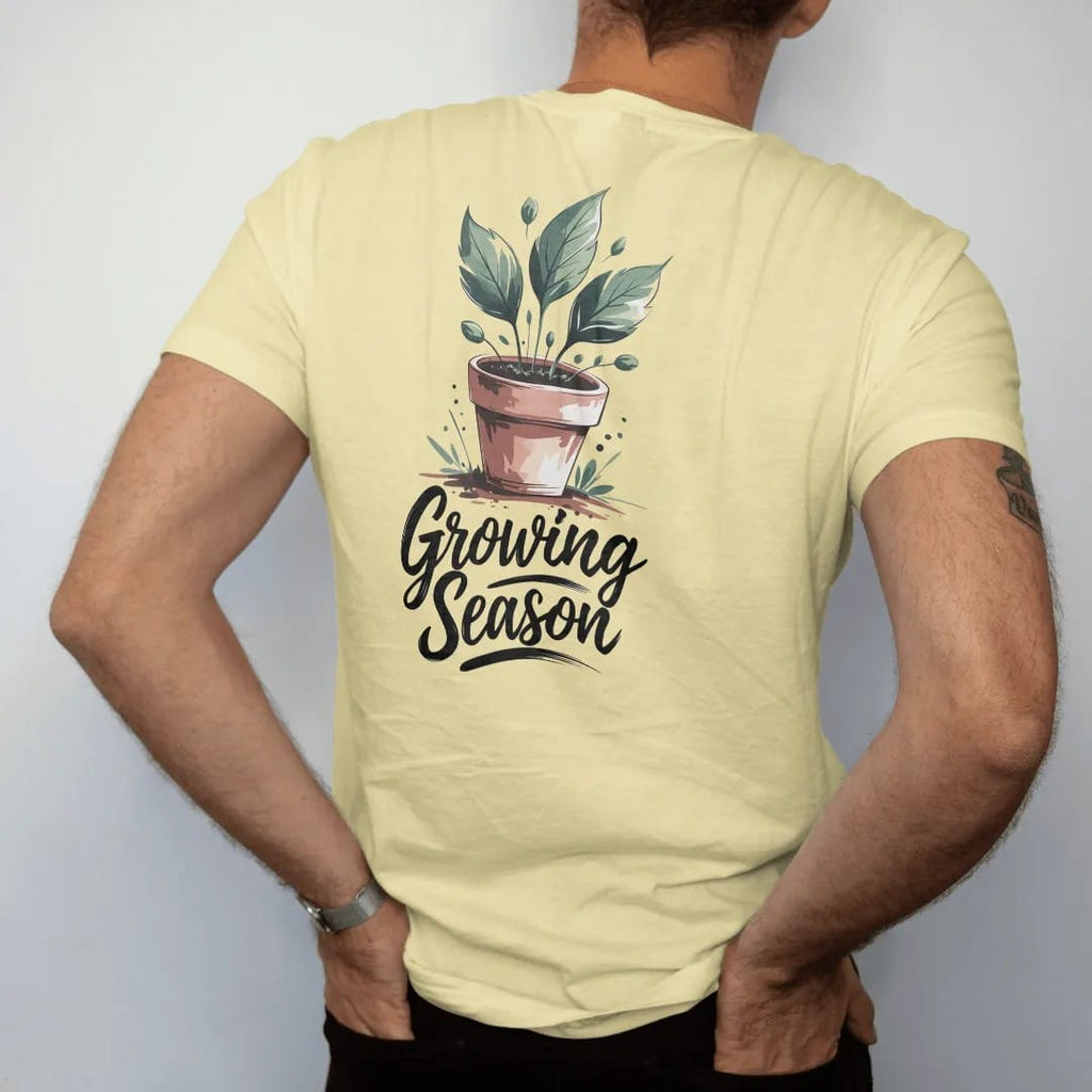 Man wearing a Butter tee, seen from the back: a small pot sprouting three fresh leaves with a few tiny buds, "GROWING SEASON" in hand-lettered brush script beneath, painted in loose watercolor and ink with soft deep green, terracotta, soil brown…