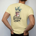 Man wearing a Butter tee, seen from the back: a small pot sprouting three fresh leaves with a few tiny buds, "GROWING SEASON" in hand-lettered brush script beneath, painted in loose watercolor and ink with soft deep green, terracotta, soil brown…
