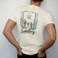 Man wearing a Ivory tee, seen from the back: a sunny windowsill with three small potted plants leaning toward a round sun, "SUNBATHING" in loose brush script beneath, painted in loose watercolor and ink with soft deep green, terracotta, soil brown…