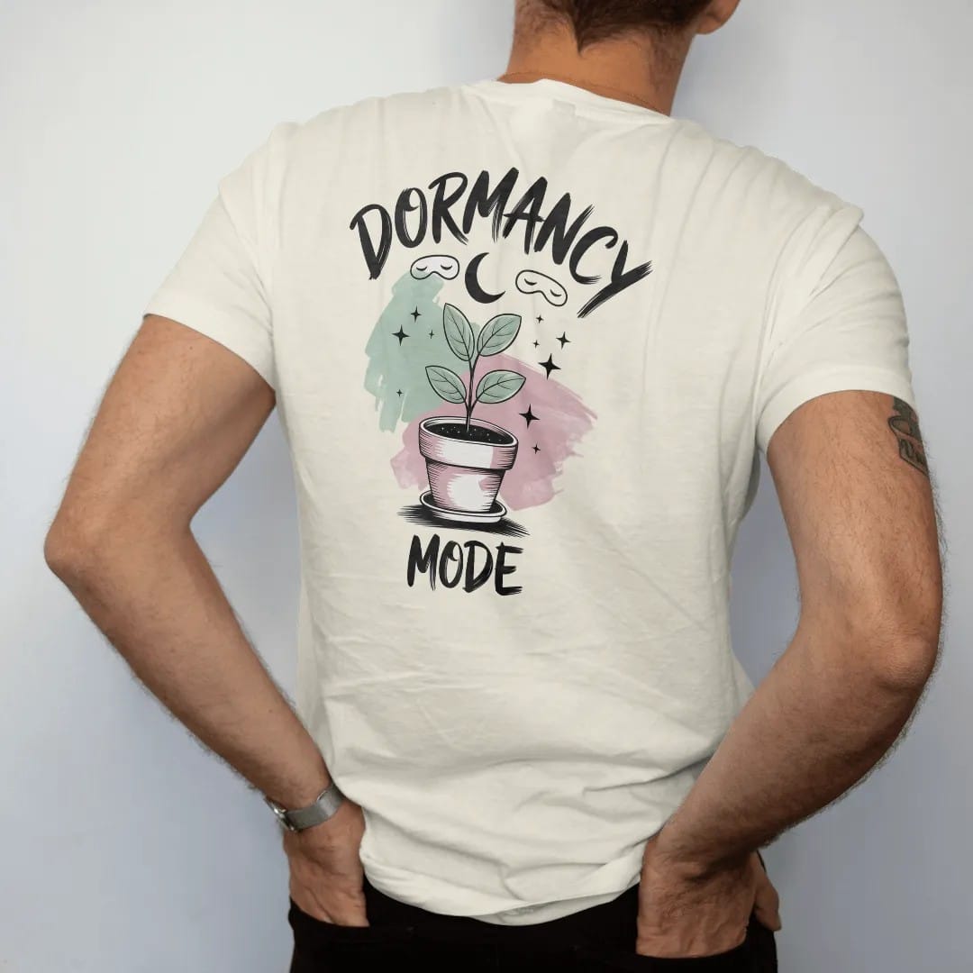 Man wearing a Ivory tee, seen from the back: a small plant in a pot with a tiny sleep mask and a crescent moon above, "DORMANCY" in loose brush script above and "MODE" small below, painted in loose watercolor and ink with soft mint, clay pink,…