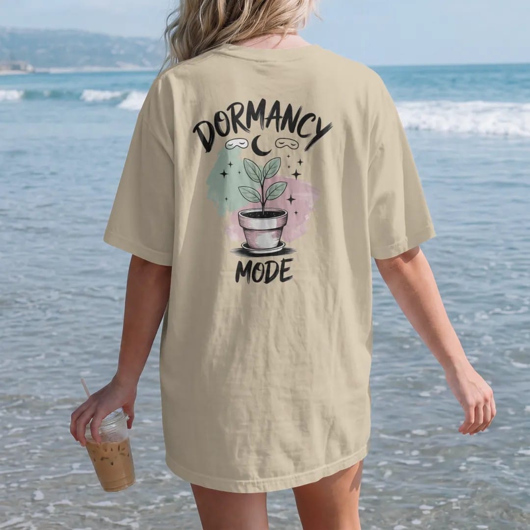 Woman on the beach in a Sandstone tee, back view: a small plant in a pot with a tiny sleep mask and a crescent moon above, "DORMANCY" in loose brush script above and "MODE" small below, painted in loose watercolor and ink with soft mint, clay pink,…
