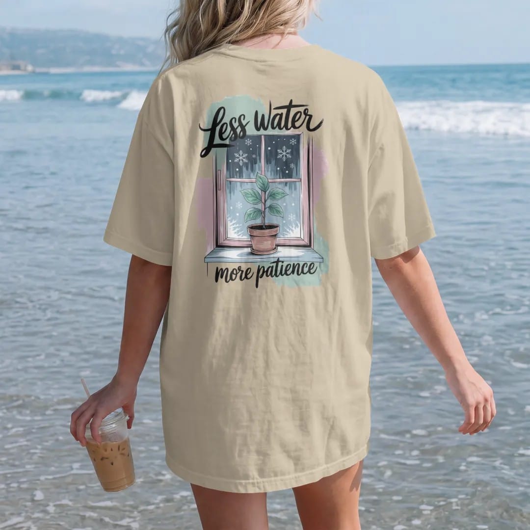 Woman on the beach in a Sandstone tee, back view: a small plant on a frosty windowsill with a few snowflakes outside the glass, "LESS WATER" in loose brush script above and "MORE PATIENCE" small below, painted in loose watercolor and ink with soft…