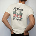 Man wearing a Ivory tee, seen from the back: three small pots in a row like a family photo, each plant a different height, "MY PLANTS" in loose brush script above and "ARE MY KIDS" below, painted in loose watercolor and ink with soft deep green,…