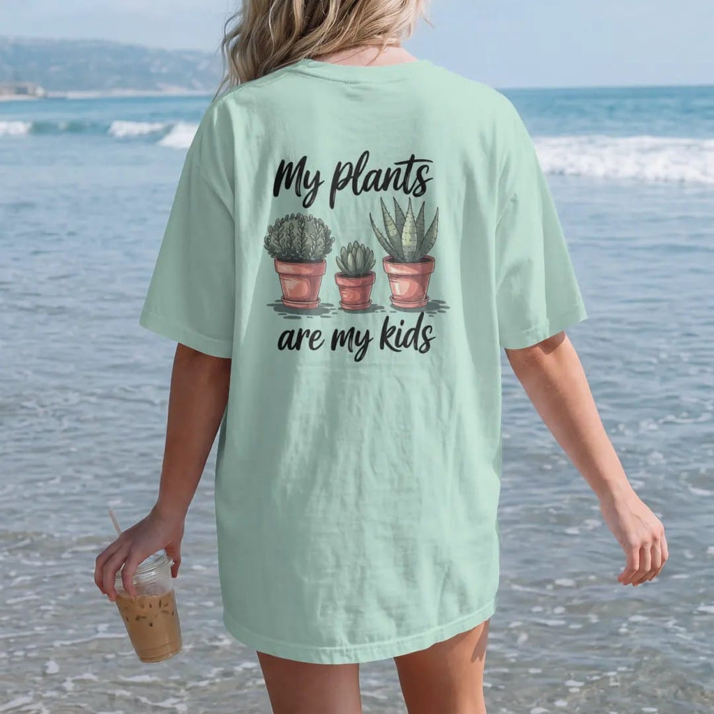 Woman on the beach in a Chalky Mint tee, back view: three small pots in a row like a family photo, each plant a different height, "MY PLANTS" in loose brush script above and "ARE MY KIDS" below, painted in loose watercolor and ink with soft deep…