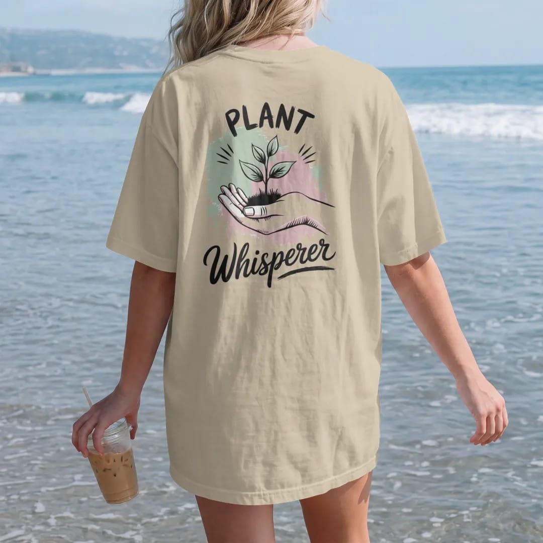 Woman on the beach in a Sandstone tee, back view: a hand cupped beside a small plant with three tiny sound lines, "PLANT" in small brush-written caps above and "WHISPERER" in hand-lettered brush script below, painted in loose watercolor and ink…