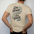 Man wearing a Sandstone tee, seen from the back: a single long trailing vine that curls slowly across the space with small leaves, "SLOW GROWTH" in loose brush script above and "IS STILL GROWTH" below, painted in loose watercolor and ink with soft…