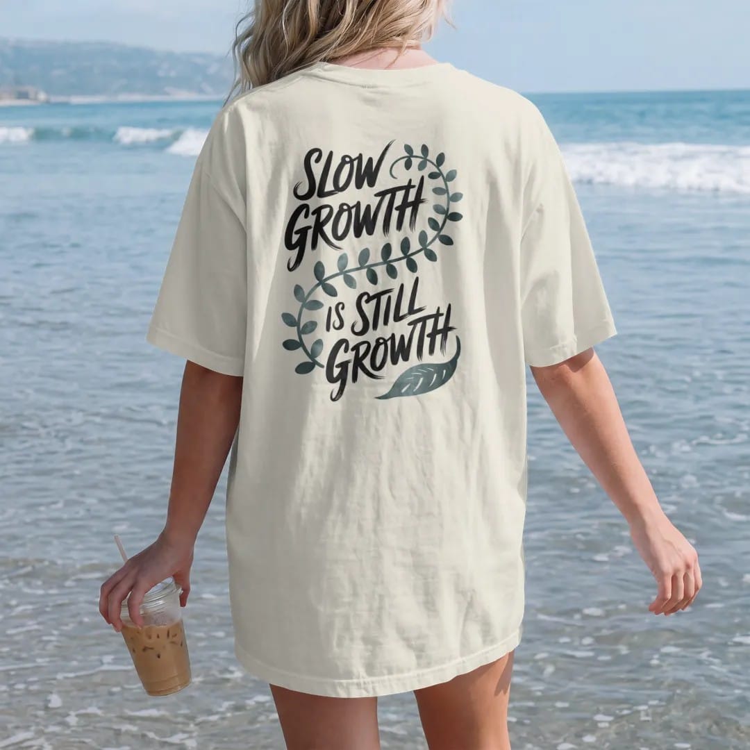 Woman on the beach in a Ivory tee, back view: a single long trailing vine that curls slowly across the space with small leaves, "SLOW GROWTH" in loose brush script above and "IS STILL GROWTH" below, painted in loose watercolor and ink with soft…