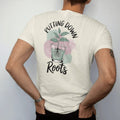 Man wearing a Ivory tee, seen from the back: a small plant in a clear glass pot showing a neat web of roots, "PUTTING DOWN" in small brush-written caps above and "ROOTS" in loose brush script below, painted in loose watercolor and ink with soft…