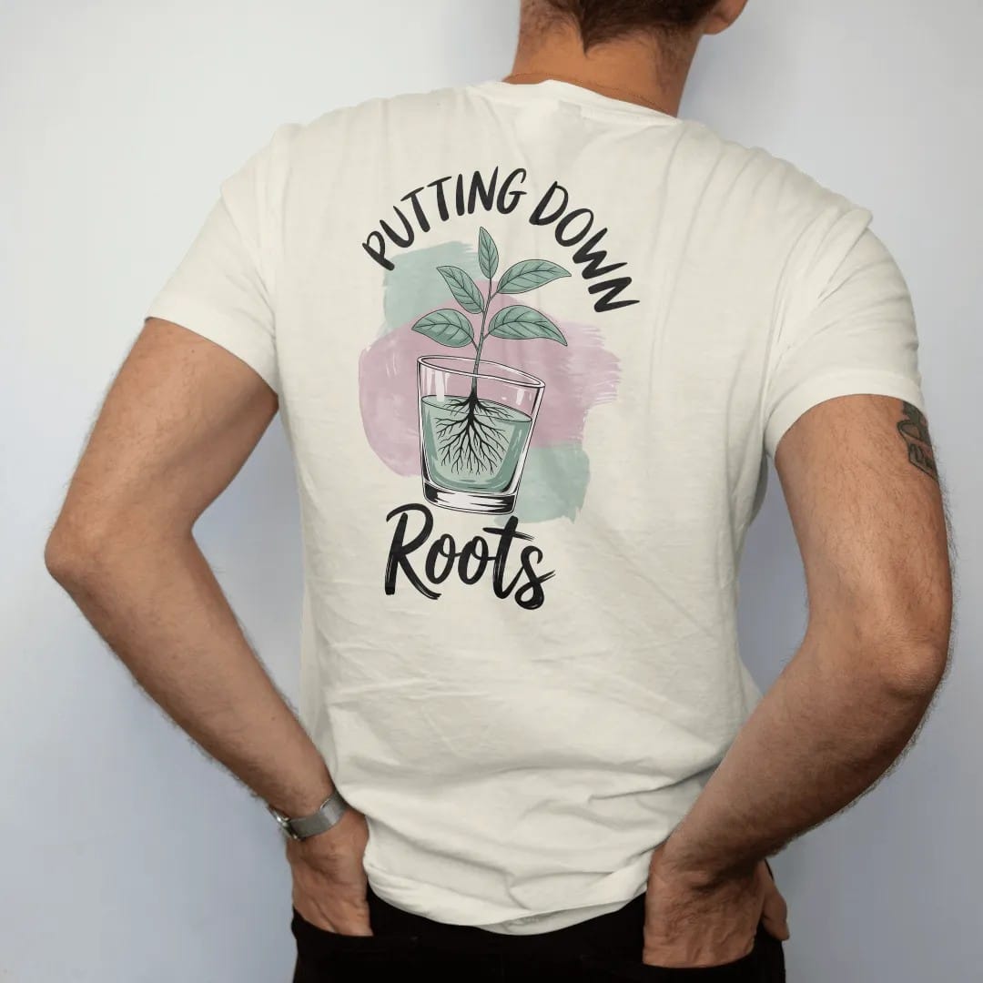 Man wearing a Ivory tee, seen from the back: a small plant in a clear glass pot showing a neat web of roots, "PUTTING DOWN" in small brush-written caps above and "ROOTS" in loose brush script below, painted in loose watercolor and ink with soft…