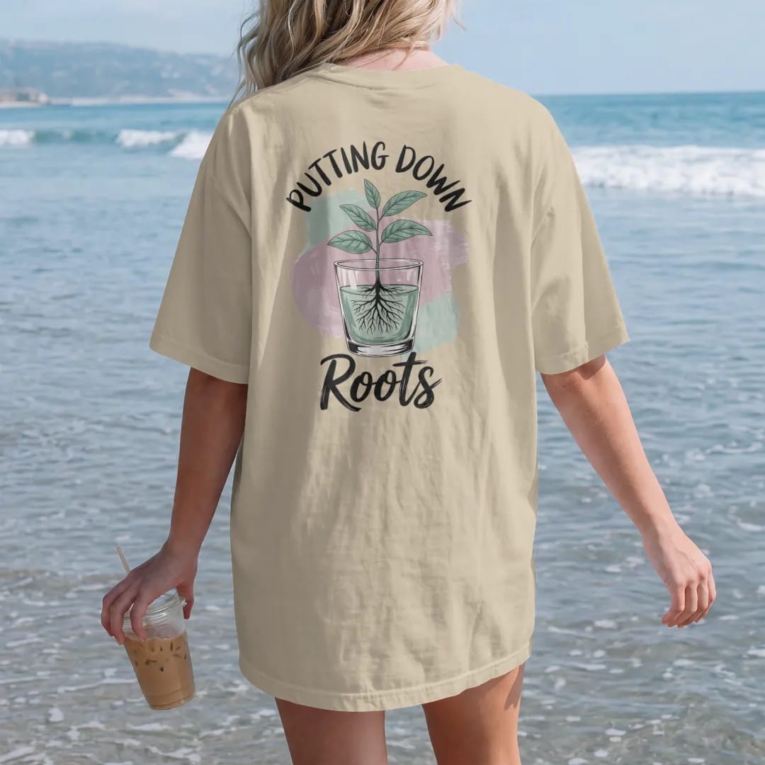 Woman on the beach in a Sandstone tee, back view: a small plant in a clear glass pot showing a neat web of roots, "PUTTING DOWN" in small brush-written caps above and "ROOTS" in loose brush script below, painted in loose watercolor and ink with…
