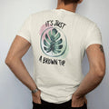 Man wearing a Ivory tee, seen from the back: a single large leaf with one small brown tip circled in a loose brush line, "IT'S JUST" in small brush-written caps above and "A BROWN TIP" in loose brush script below, painted in loose watercolor and…