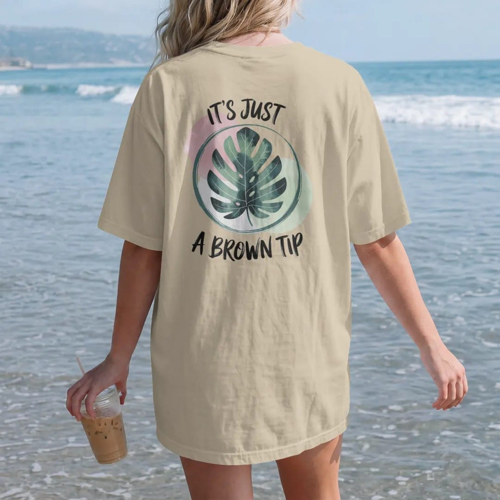 Woman on the beach in a Sandstone tee, back view: a single large leaf with one small brown tip circled in a loose brush line, "IT'S JUST" in small brush-written caps above and "A BROWN TIP" in loose brush script below, painted in loose watercolor…