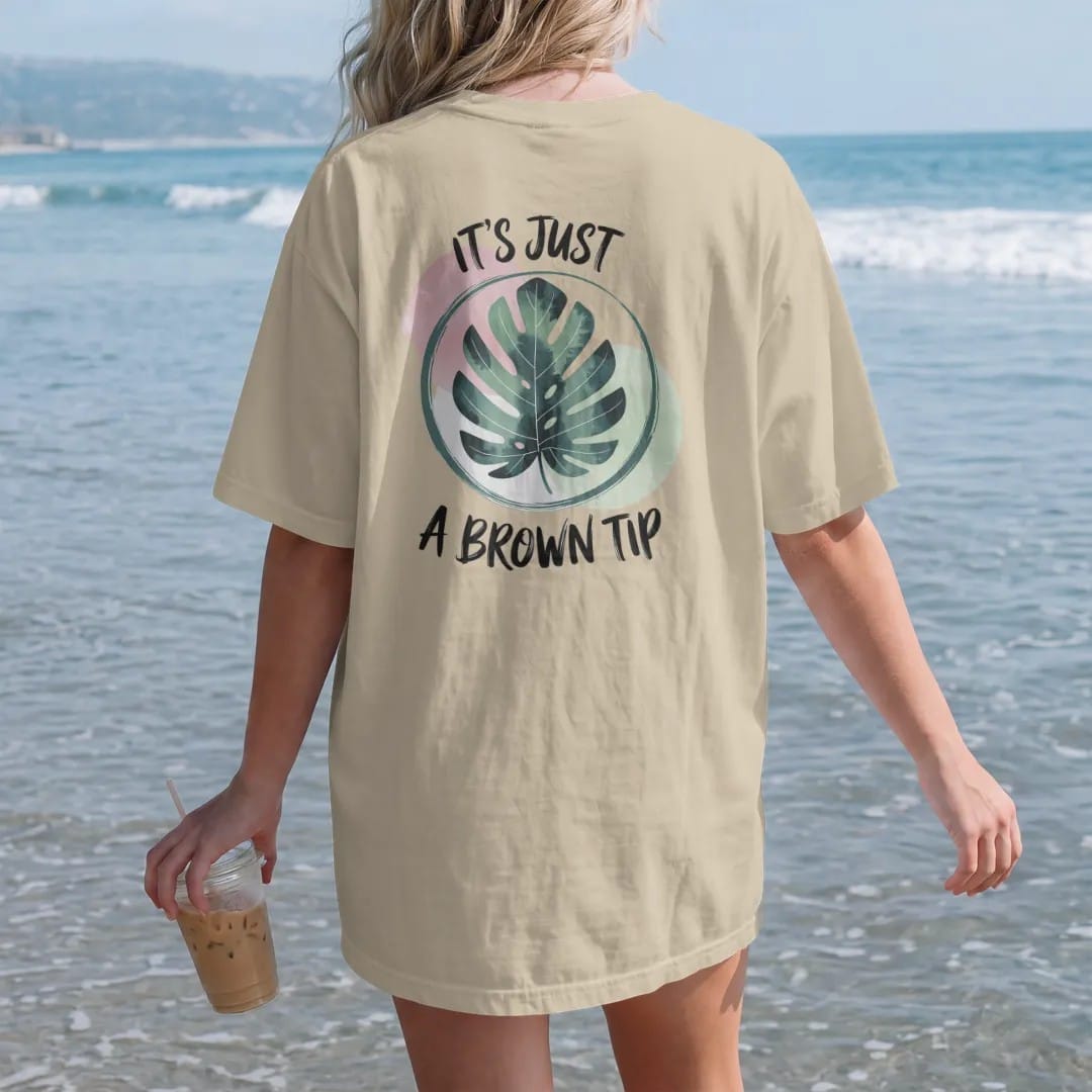 Woman on the beach in a Sandstone tee, back view: a single large leaf with one small brown tip circled in a loose brush line, "IT'S JUST" in small brush-written caps above and "A BROWN TIP" in loose brush script below, painted in loose watercolor…
