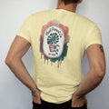 Man wearing a Butter tee, seen from the back: a neat oval crest with a monstera leaf and a pothos vine framing a small pot, "PLANT PARENT CO." in loose brush script above and "EST. 2026" below, painted in loose watercolor and ink with soft deep…